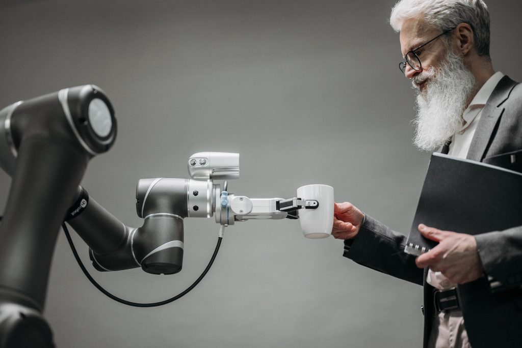 Ein Roboterarm übergibt eine weiße Tasse an einen Mann mit Brille und grauem Bart in einem neutralen Raum.